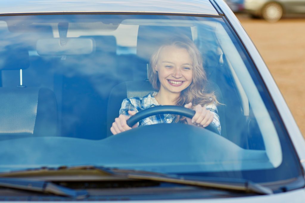 มองจากด้านนอกรถ ผู้หญิงผมทองนั่งจับพวงมาลัยขับรถ ยิ้มมีความสุข